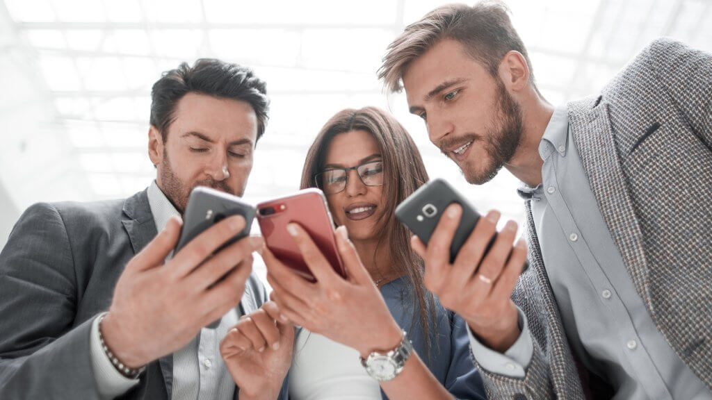 Three young work colleagues stood together, looking at their phones and smiling
